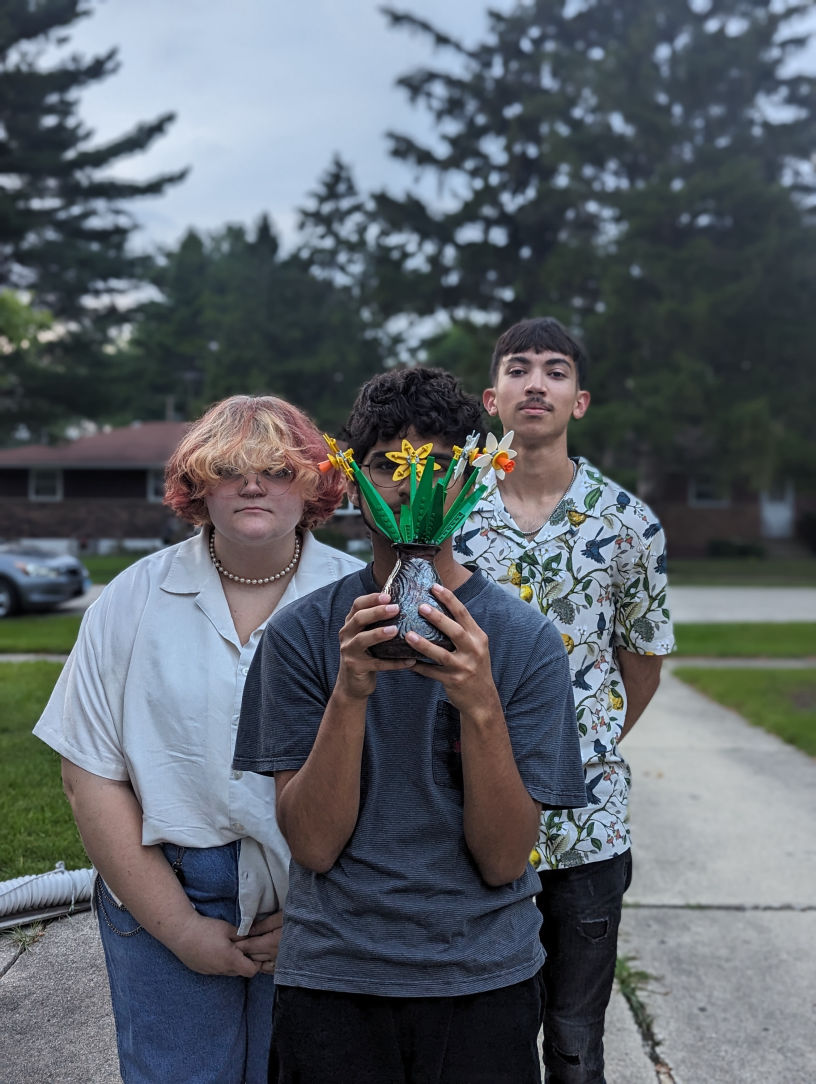 A photo of the band, with Adrian standing in front of the camera holding lego flowers, and Alexie and Os standing behind him.